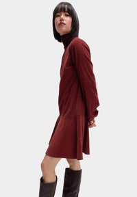 Bordowa sukienka z długim rękawem, z wysokim kołnierzem, luźnym krojem, detalami panelu bocznego i rozkloszowanym dołem. Noszona z brązowymi, do kolan, botkami.
