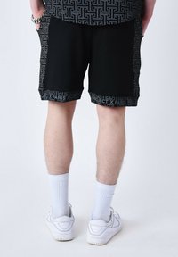Shorts noirs avec un motif géométrique, arborant une bande logo au niveau de l'ourlet. Portés avec des chaussettes blanches et des baskets, affichant un style décontracté et sportif.