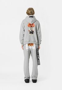 Sudadera gris con un gráfico de un oso de peluche en la parte trasera y pantalones de chándal a juego con texto en negrita y un detalle de etiqueta en el lateral. Calzado negro incluido.
