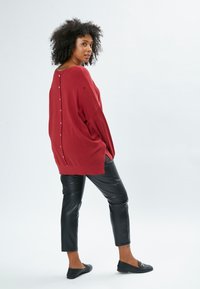 Übergroßer bordeauxroter Pullover mit V-Ausschnitt und versteckten Knöpfen auf der Rückseite, kombiniert mit schwarzen Lederhosen und flachen Schuhen. Angenehme Textur.