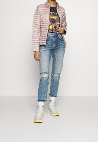 Light pink puffer jacket with a high collar, paired with a graphic black t-shirt, distressed blue jeans, and gray sneakers with yellow accents.