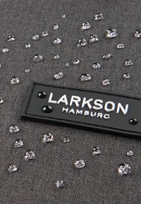 Grått tyg med glittrande ädelstenar och en svart LARKSON HAMBURG-etikett. Texturen är slät med en subtil lyster.