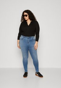 Μαύρο μακρυμάνικο πόλο μπλουζάκι, ανοιχτό μπλε skinny τζιν, μαύρα αθλητικά παπούτσια με λευκές λεπτομέρειες και μεγάλα γυαλιά ηλίου. Το φόντο είναι απλό γκρι.