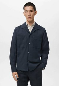 Chaqueta azul marino abotonada, fabricada con tela texturizada, que presenta un cuello, dos bolsillos en el pecho y un corte recto. Llevada sobre una camisa clara.