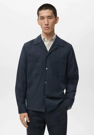 Chaqueta azul marino abotonada, fabricada con tela texturizada, que presenta un cuello, dos bolsillos en el pecho y un corte recto. Llevada sobre una camisa clara.