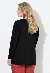 Schwarzes Langarmoberteil mit rundem Halsausschnitt, kombiniert mit glänzenden roten Hosen. Blonde Haare fallen in Wellen über die Schulter. Rote Ohrringe betonen den Look.