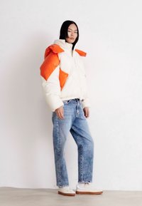 Giubbotto imbottito bianco e arancione con zip e cappuccio, caratterizzato da un design geometrico. Abbinato a jeans in denim blu chiaro e scarpe bianche.