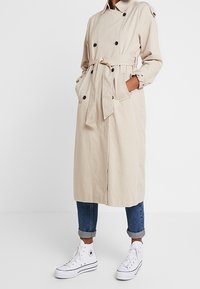 Trench coat beige con bottoni doppi, vita con cintura e tasche laterali; indossato sopra jeans di denim arrotolati e sneakers bianche.