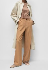 Beżowy płaszcz trenczowy na brązowej dopasowanej bluzce, szerokie beżowe spodnie i jasnoróżowe baletki z krzyżującymi się paskami. Gładkie wykończenie tkaniny.