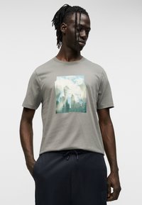Γκρι κοντομάνικο μπλουζάκι με ορθογώνιο τύπωμα που απεικονίζει βουνά και δέντρα σε αποχρώσεις μπλε και πράσινου. Άνετη εφαρμογή, στρογγυλή λαιμόκοψη.