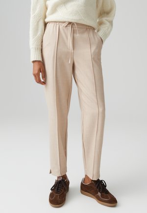 Helle beige, plissierte Hose mit elastischem Bund und seitlichen Taschen, kombiniert mit braunen Wildledersneakern. Glatte Stofftextur.