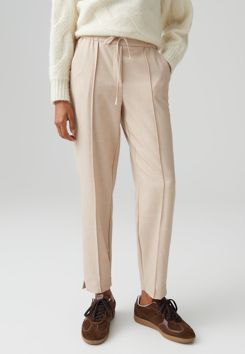 Lichtbeige geplooide broeken met een elastische tailleband en zijzakken, gecombineerd met bruine suède sneakers. Gladde stofstructuur.