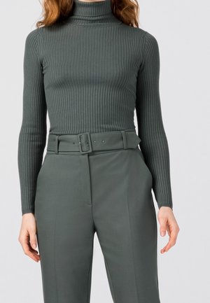 Femme portant un pull col roulé ajusté à côtes et un pantalon taille haute ceinturé, tous deux dans des tons de vert-gris en sourdine.
