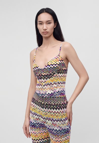 Jeune femme aux longs cheveux foncés portant une combinaison sans manches à motif zigzag coloré sur un fond clair uni.