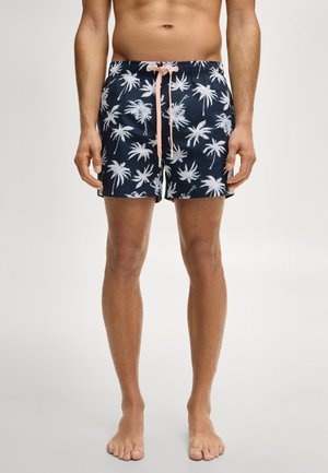 Torso et jambes d'un homme vêtu d'un short de bain bleu marine orné d'imprimés de palmiers blancs et d'un cordon de serrage rose clair, debout pieds nus sur un fond uni.