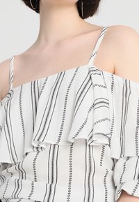 Top bianco con design scollo a spalle scoperte, caratterizzato da strisce verticali nere e dettagli arricciati. Tessuto liscio e leggero.