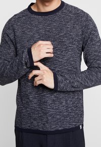 Man som bär en marinblå och vit spräcklig stickad tröja och justerar manschetten på sin vänstra ärm, sedd från axlarna till midjan.