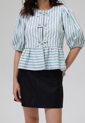 Femme portant un chemisier à rayures verticales vertes et blanches avec des manches bouffantes et des nœuds, associé à une jupe noire.