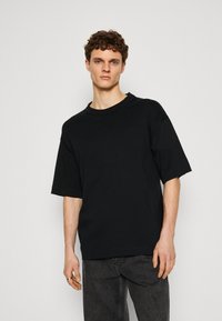 T-shirt nera in cotone oversize con colletto rotondo e maniche corte, caratterizzata da un design minimalista e priva di loghi o motivi visibili.