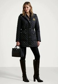 Sieviete, kas valkā melnu quiltētu mēteļa ar kapuci, melnas legingu, melnas augstas zābaku līdz ceļgaliem un turēt melnu rokassomu.