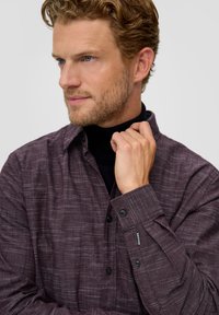 Langärmliges Hemd in dunklem Kastanienbraun mit dezenten horizontalen Streifen, Button-Down-Kragen und schwarzen Knöpfen; über einem schwarzen Rollkragenpullover getragen.