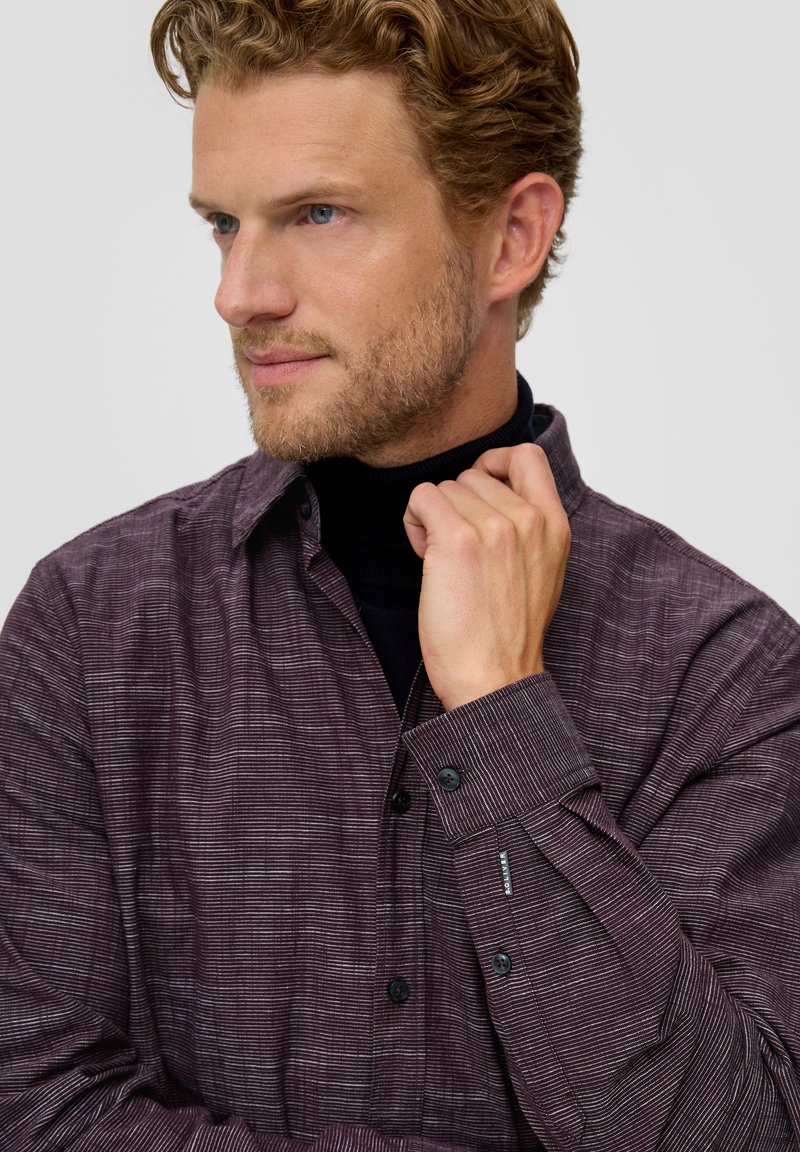 Camisa de manga larga en marrón oscuro con sutiles rayas horizontales, collar abotonado y botones negros; llevada sobre un cuello de tortuga negro.