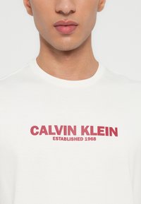 Άνδρας που φοράει λευκό T-shirt με λαιμόκοψη, στο οποίο είναι τυπωμένο το κόκκινο "CALVIN KLEIN ESTABLISHED 1968" στο στήθος.