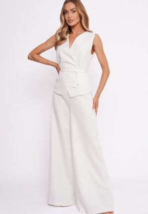 Kobieta stojąca z ręką na biodrze, ubrana w bezrękawnikowy biały top z kopertowym dekoltem i paskiem oraz szerokie białe spodnie na tle jednolitego tła.