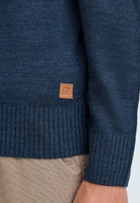 Šviesiai mėlynas megztinis su mėgintuvėlio raštu, su šonkauliniais rankogaliais ir apačia, ant apatinės dalies esantis mažas odinis etiketė su prekės ženklo logotipu.