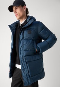 Giacca imbottita blu navy con cappuccio, caratterizzata da una zip frontale, tasche laterali e una patch con logo sul petto. Texture liscia e trapuntata.