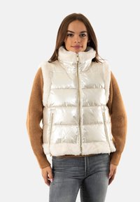 Gilet argentato metallico voluminoso con rifiniture e colletto in pelliccia sintetica, chiusura con zip sul davanti e tasche laterali, indossato sopra un maglione marrone.