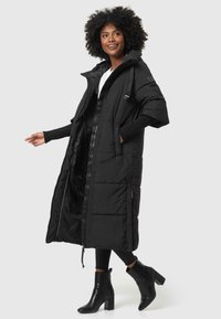 Lang sort pufferjakke med hætte, lynlås foran, quiltet design, sidelommer og ribbede strikmanchetter, båret over mørke jeans og sorte hælboote.