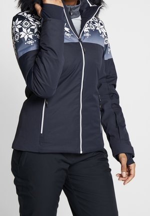 Femme portant une veste d'hiver bleu marine avec des motifs de flocons de neige blancs sur les épaules et une capuche bordée de fourrure.