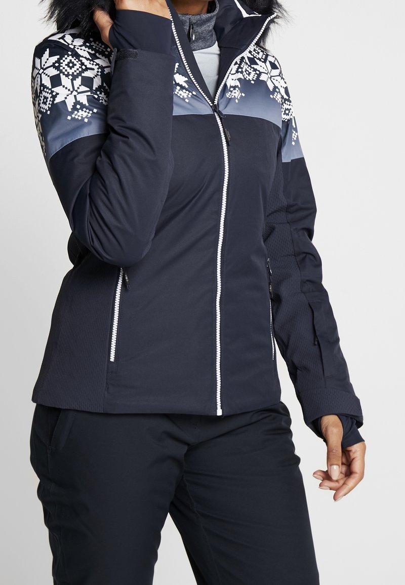 Femme portant une veste d'hiver bleu marine avec des motifs de flocons de neige blancs sur les épaules et une capuche bordée de fourrure.