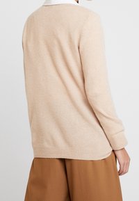 Suéter beige claro fabricado con material suave. Presenta puños y dobladillo de punto acanalado, un corte ligeramente holgado y un diseño simple en la parte posterior.