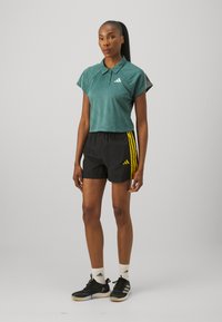 Teal geknipt polo shirt met korte mouwen, gecombineerd met zwarte shorts met gele strepen en het Adidas-logo; zwarte sportschoenen en witte sokken.