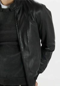 Giacca di pelle nera con finitura liscia, dotata di colletto alto e chiusura con zip. Indossata sopra un maglione grigio scuro a texture.