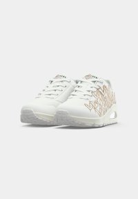 White sneakers with a textured mesh design, featuring a slight heel and decorative beige stitching in a heart pattern. Rubber outsole.