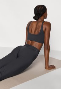 Top sportivo nero e leggings a vita alta in tessuto elasticizzato, indossati da una persona in posizione inclinata su un tappetino da yoga beige.