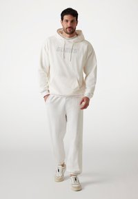 Hellbeige Kapuzenpullover mit geripptem Textur und Fronttasche, kombiniert mit passenden Jogginghosen und weißen Sneakers.