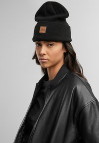 Berretto di maglia nera con parte superiore sformata, dotato di una toppa in pelle quadrata etichettata "URBAN CLASSICS." Indossato con una giacca bomber in pelle nera.