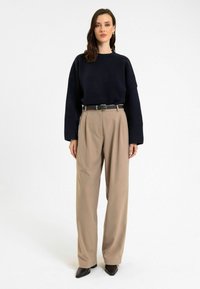 Donkerblauwe gebreide trui met lange mouwen, gecombineerd met beige hoge taille broek en een zwarte riem. Zwarte schoenen met een spits toelopende neus.