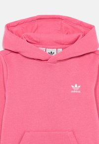 Sweatshirt rosa feito de algodão suave, com um bolso frontal, capuz com cordão e um logo Adidas branco no peito. Textura suave.