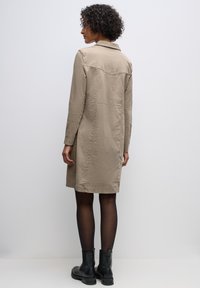 Beige dlouhý rukáv šaty s límečkem a detaily švu na zadní straně, doplněné černými kotníkovými botami, zobrazené zezadu na neutrálním pozadí.