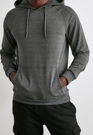 Mann trägt einen mittelgrauen Kapuzenpullover mit Fronttasche und schwarze Hose, Hände in den Taschen, vor einfachem Hintergrund.