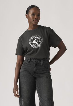 Femme portant un t-shirt noir avec un motif de boule à facettes blanche, associé à un jean foncé taille haute, posant devant un fond uni.