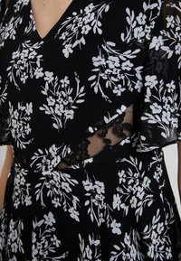 Mustassa mekossa on valkoinen kukkakuosi, jossa on läpinäkyviä pitsiyksityiskohtia ja lyhyet hihat. V-kaula-aukko, kapea vyötärö ja hulmuava hame.