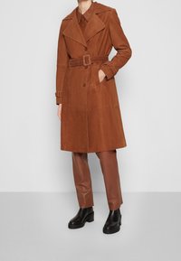 Abrigo tipo gabardina de ante marrón con solapas anchas, cintura con cinturón y botones de doble fila. Modelo con pantalones de cuero marrón y botas negras.