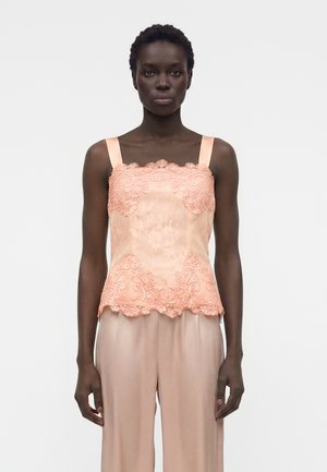 Femme portant un caraco en dentelle couleur pêche avec de larges bretelles et un pantalon soyeux assorti, debout devant un fond blanc uni.