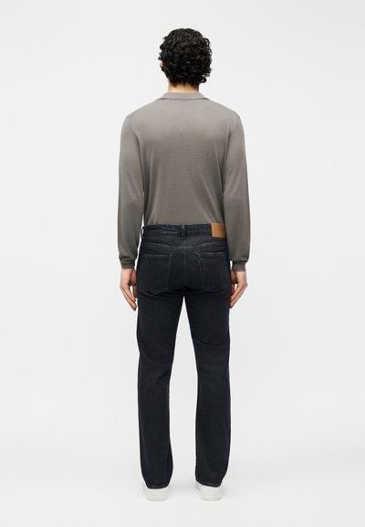 Άνδρας με σγουρά μαλλιά που στέκεται πλάτη προς το φακό, φορώντας γκρι μπλούζα με μακριά μανίκια, μαύρο τζιν και λευκά αθλητικά παπούτσια, μπροστά σε απλό φόντο.